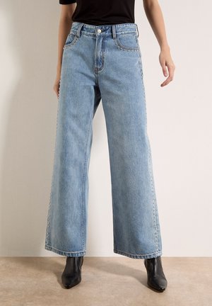 Jeans azzurri a gamba larga con borchie metalliche lungo le tasche e le cuciture esterne, indossati con stivaletti neri con punta appuntita.