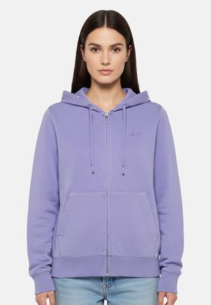 Donna con capelli lisci e scuri che indossa una felpa con cappuccio color lilla chiaro con cerniera, tasche frontali e coulisse, abbinata a jeans azzurri.