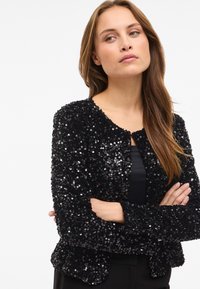 Črna jakna s sequini, okroglo izrezana in z dolgimi rokavi. Ima teksturirano površino, ki jo tvorijo tesno razporejeni sequini.