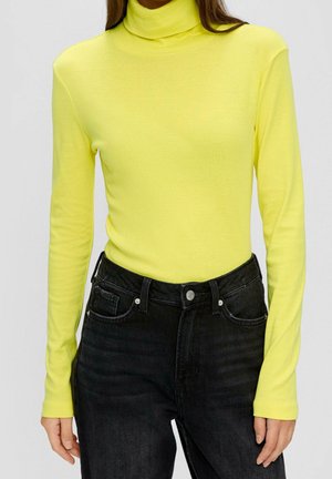 Femme portant un haut à col roulé jaune vif à manches longues, rentré dans un jean noir taille haute, debout les bras relâchés.