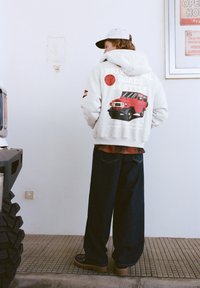 Šedá mikina s kapucí s červeným potiskem starého nákladního auta, doplněná tmavými širokými džínami a černými botami, pohled zezadu.