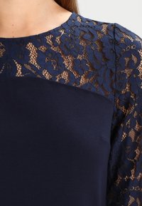 Haut bleu marine avec manches en dentelle à motif floral, partie du corps lisse, décolleté rond, et bordure en dentelle texturée.