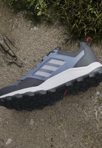 Baskets Adidas grises et bleues avec une semelle noire rugueuse reposant sur un chemin de gravier à côté d'herbe verte et de plantes.