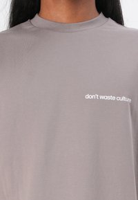 Camiseta gris con cuello redondo y mangas cortas, que presenta el texto "no desperdicies la cultura" impreso en blanco en el frente.