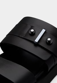 Sandales en cuir noir avec deux larges brides, design élégant. Présentent des accents en métal noir et une pièce métallique distinctive sur la bride.