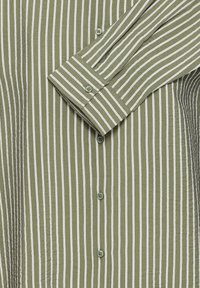 Camicia verde con bottoni e strisce bianche verticali, con il polsino e la manica piegata sulla parte anteriore.