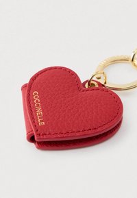 Breloc în formă de inimă din piele texturată roșie, cu logo-ul auriu Coccinelle și inel metalic auriu pe un fundal alb.