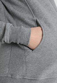 Mano inserita nella tasca anteriore di un felpa grigia a maglia con polsini a coste e dettagli di cucitura visibili.
