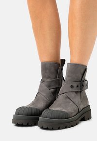 Šedé kotníkové boty s texturovaným koženým svrškem, gumovou špičkou, vrstveným detailem pásku, sponou a robustní podrážkou.