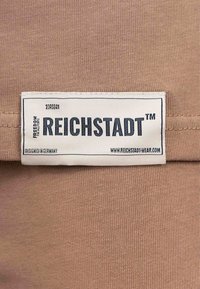 Tessuto color sabbia con una trama particolare, dotato di un'etichetta bianca con "REICHSTADT™" in grassetto, insieme ai dettagli del marchio sottostanti.
