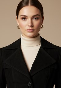 Jonge vrouw met bruin haar naar achteren gebonden, draagt gouden oorringen, een crèmekleurige coltrui en een zwarte jas met een grote revers.