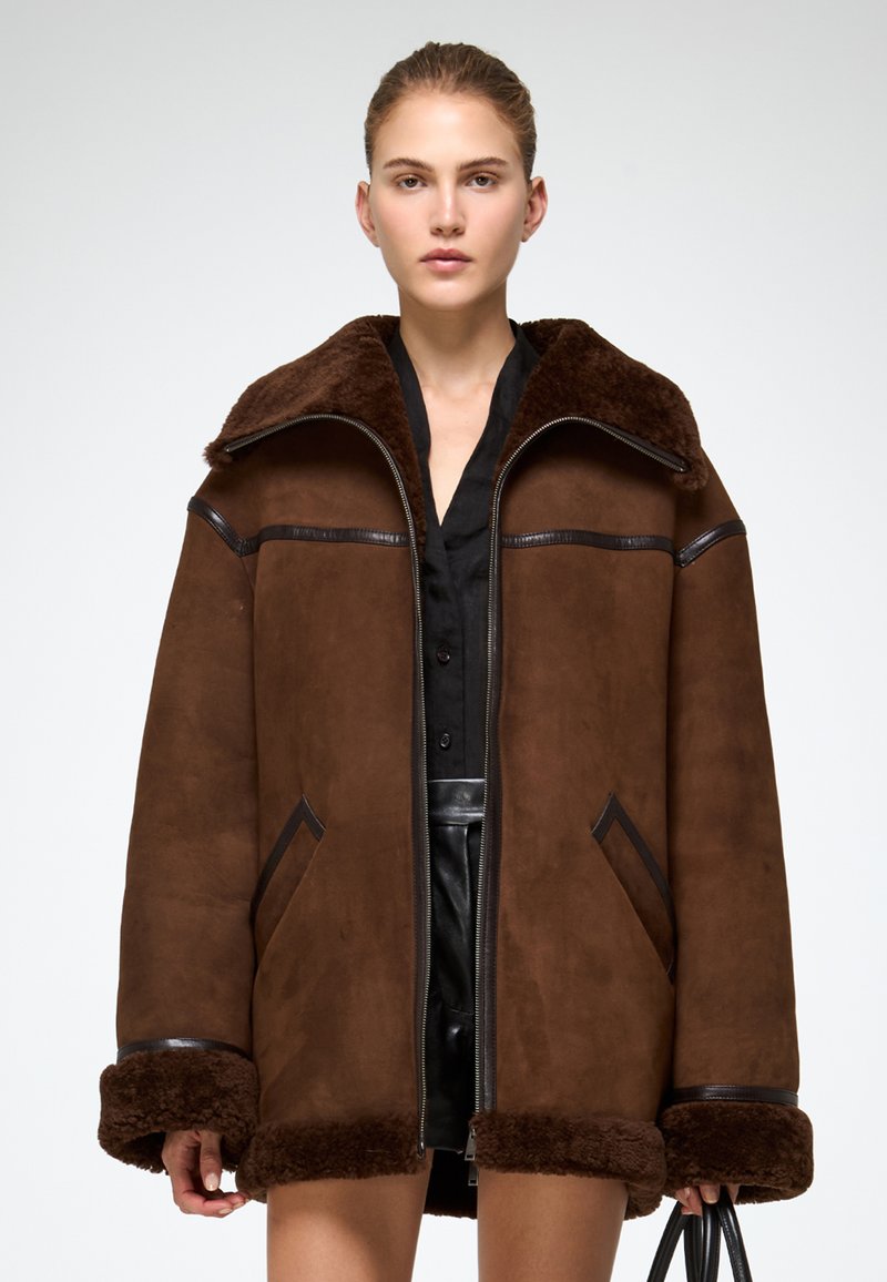 Pruunist suedest jope, millel on karvast vooderdatud krae, naha aktsendid ja kaks esikotti. Sisaldab tõmblukuga kinnitamist ja laiu mansette.