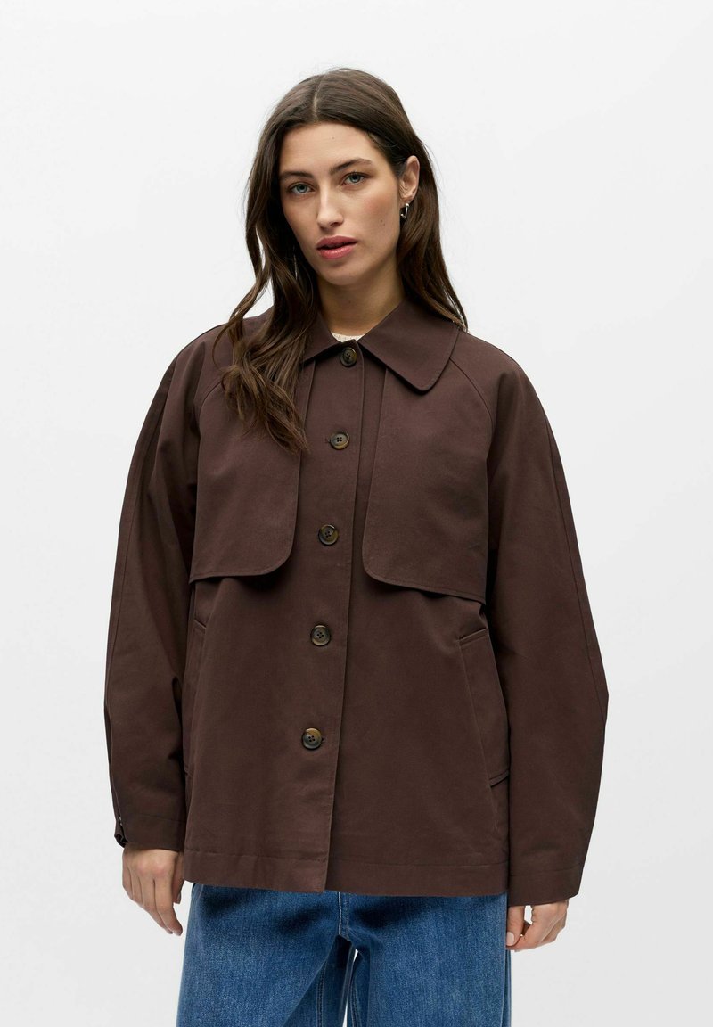 Femme aux longs cheveux bruns portant une veste marron foncé boutonnée avec un détail à rabat, sur un jean bleu, debout devant un fond blanc.