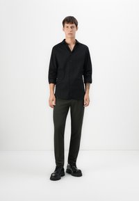 Joven vestido con una camisa negra de botones, pantalones verde oscuro y zapatos chunky negros, de pie contra una pared blanca sencilla.