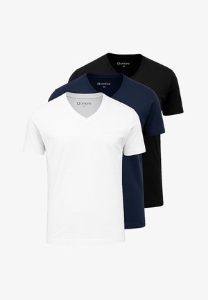 Mand iført sort V-hals t-shirt, grå ternede bukser, hvide sneakers, briller og et armbåndsur, der står med hænderne i lommerne.