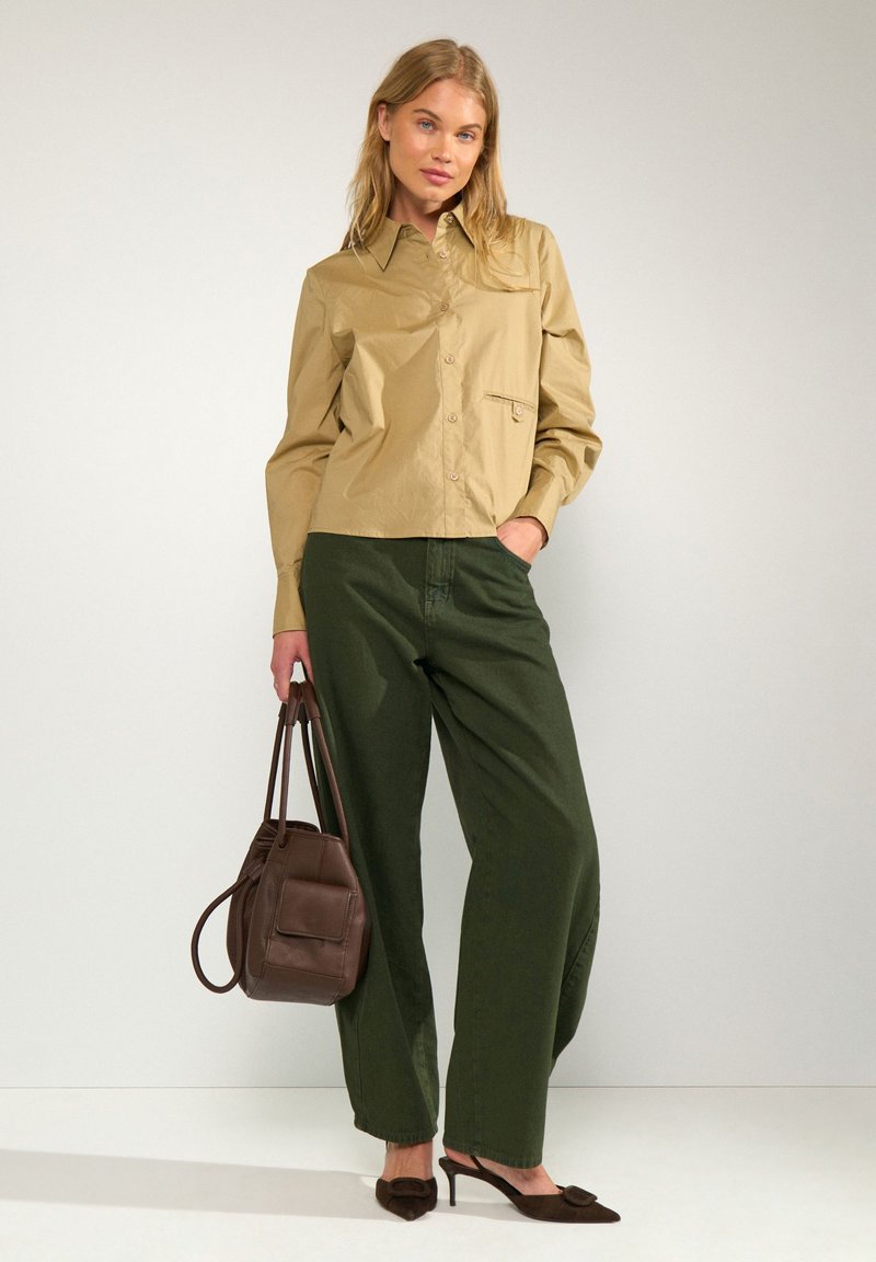 Femme portant une chemise beige boutonnée, un pantalon large vert foncé, des talons pointus marron foncé et tenant un sac à main marron foncé.