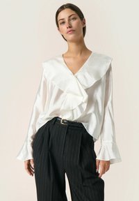 Blusa de satén blanca con un escote en V profundo y un gran cuello con volantes, combinada con pantalones negros a rayas con cintura ceñida.