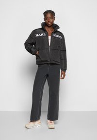 Giacca a vento nera con colletto alto e branding bianco "KARL" sul petto, abbinata a jeans scuri e sneakers beige.