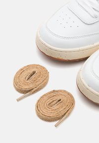 White leather sneakers with perforated toe and beige rubber sole. Two rolls of tan jute laces with a cardboard tip laid beside the shoes.