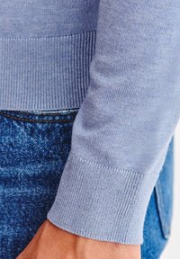 Jasnoniebieski sweter z dzianiny z prążkowanymi mankietami, prezentujący gładką teksturę. Noszony z ciemnymi dżinsami, ukazujący zbliżenie detali rękawa.
