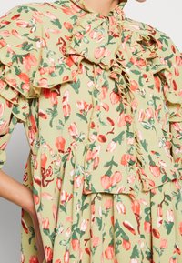 Frau in einem hellgrünen, blumenbedruckten Kleid mit roten und weißen Blumen, das mit Rüschen und einer großen Schleife am Ausschnitt versehen ist.