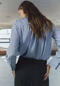 Blusa azzurro chiaro a maniche lunghe con vestibilità morbida e sottili righe verticali, abbinata a pantaloni scuri su misura con una texture liscia.