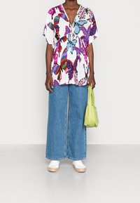 Persona che indossa una blusa ampia a stampa farfalla colorata, jeans larghi blu, scarpe platform bianche, mentre tiene una borsa verde chiaro su uno sfondo bianco.