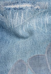 Tela de mezclilla de color azul claro con bordes deshilachados y costuras blancas, con un parche floral texturizado en un tono de azul ligeramente más oscuro.