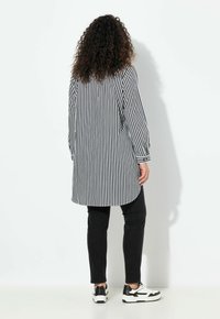 Camicia lunga a righe bianche e nere con polsini con bottoni, abbinata a pantaloni neri e sneakers bianche. Tessuto liscio, vestibilità comoda.
