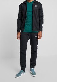 Black athletic jacket with white stripes and logo, green t-shirt, black pants with white accents, and teal sneakers with white soles.