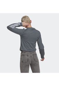 Grå långärmad skjorta med vit tre-stripes design på ärmarna, ihop med högmidjade blekta svarta jeans, som visar en elegant, avslappnad look.