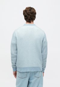 Oseba s kratkimi kodrastimi lasmi, oblečena v svetlo modro pleteno pulover in svetlo modre kavbojke, stoji obrnjena stran od preprostega belega ozadja.