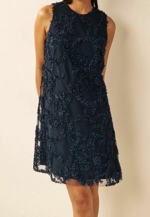 Granatowa sukienka bez rękawów z kwiatową aplikacją o fakturze, do kolan, noszona przez osobę na jasnym, jednolitym tle.