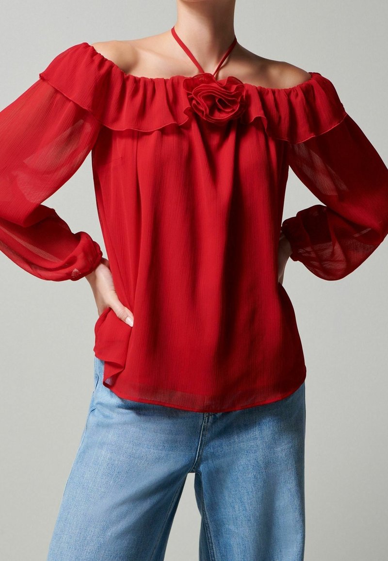 Femme portant un chemisier rouge transparent à épaules dénudées avec des volants et un détail rose, associé à un jean bleu clair, sur un fond uni.