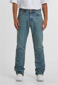 Šviesiai mėlyni džinsai tiesiu kirpimu, su kišenėmis, diržo kilpomis ir išblukusiu skalbimu su matomomis siūlėmis. Dėvimi su baltais sportbačiais.