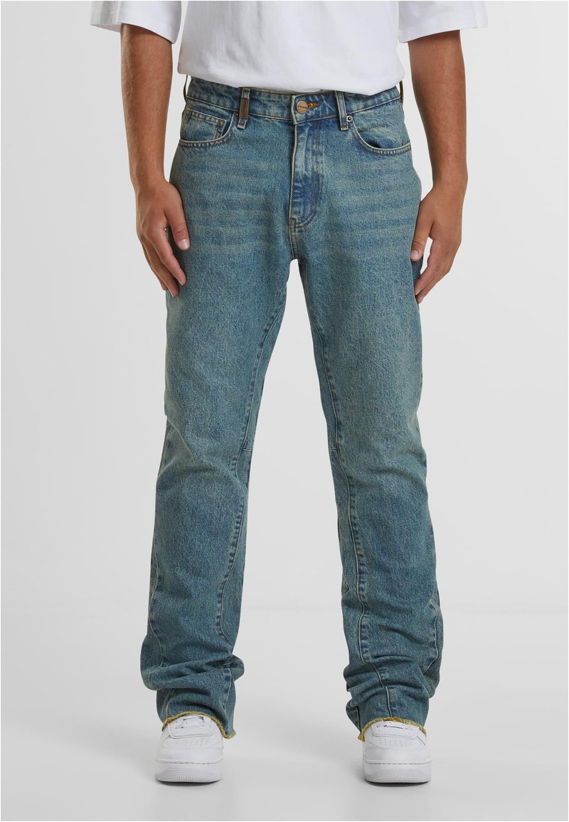 Šviesiai mėlyni džinsai tiesiu kirpimu, su kišenėmis, diržo kilpomis ir išblukusiu skalbimu su matomomis siūlėmis. Dėvimi su baltais sportbačiais.