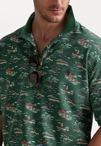 Zaļš polo krekls ar teksturētu audumu, ko rotā ainavas dizains ar ēkām un laivām, kombinēts ar tumšām saulesbrillēm.