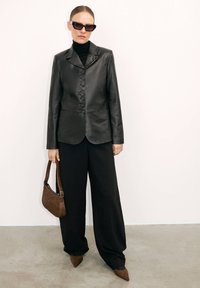 Blazer en cuir noir avec une coupe ajustée et fermeture à boutons, associé à un pantalon noir à larges jambes, un sac à main en cuir texturé marron et des ballerines pointues.
