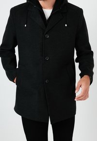 Cappotto in lana nera con doppia zip e chiusura a bottone, maniche lunghe, tasche e cappuccio con lacci regolabili. Tessuto liscio.