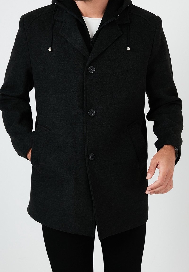 Cappotto in lana nera con doppia zip e chiusura a bottone, maniche lunghe, tasche e cappuccio con lacci regolabili. Tessuto liscio.