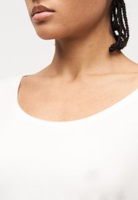 Κοντινό πλάνο του κάτω μέρους του προσώπου μιας γυναίκας, του λαιμού και του άνω μέρους του στήθους που φοράει λευκό μπλουζά κι με στρογγυλή λαιμόκοψη, με πλεξούδες στα μαλλιά, πάνω σε απλό φόντο.