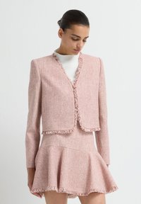 Chaqueta de tweed rosa con bordes deshilachados, combinada con una falda acampanada a juego. Textura suave, diseño estructurado y sutil detalle de patrón.
