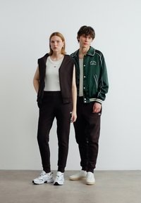 Νέος άνδρας και γυναίκα στέκονται δίπλα δίπλα φορώντας άνετα αθλητικά ρούχα με σκούρα παντελόνια, λευκά τοπ και αθλητικά παπούτσια μπροστά σε απλό λευκό τοίχο.