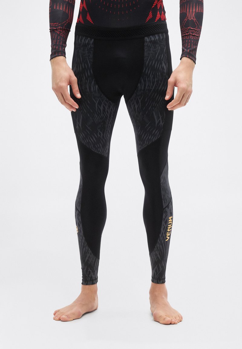 Άνδρας που φοράει μαύρο και σκούρο γκρι κολάν συμπίεσης με σχέδιο και το λογότυπο "VENUM" στο κάτω μέρος του ποδιού, όρθιος ξυπόλητος σε λευκό φόντο.