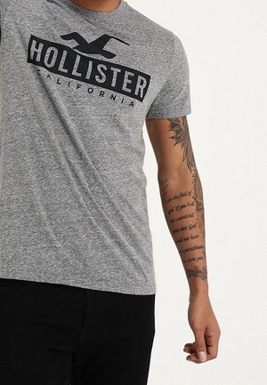 Camiseta de algodón gris con un logo negro "HOLLISTER CALIFORNIA" en el pecho. Diseño de cuello redondo y mangas cortas.