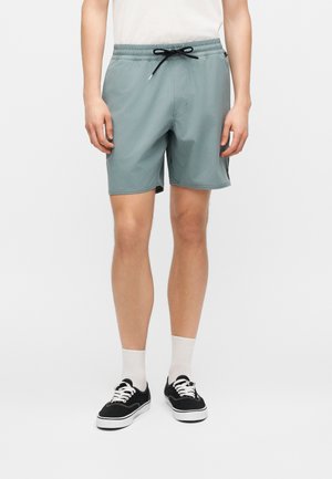 Mand iført lysegrå elastiske shorts med sort snøre, hvide sokker og sorte lave sneakers på ensfarvet baggrund.