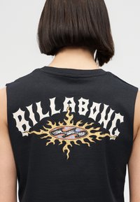 Άτομο με κοντά καστανά μαλλιά που φοράει μαύρο μπλουζάκι χωρίς μανίκια με γραφικό φλεγόμενου ήλιου και το κείμενο «Billabong» στην πλάτη.