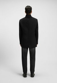 Cappotto nero con colletto alto, taglio dritto e texture liscia. Abbinato a jeans scuri e scarpe nere robuste, visto da dietro.