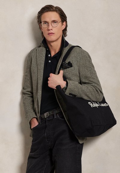 Νέος άνδρας με γυαλιά φορώντας σακάκι με σχέδιο ψαροκόκαλο, μαύρο πόλο πουκάμισο, σκούρα τζιν, κρατώντας μια μαύρη τσάντα tote Ralph Lauren στον ώμο.