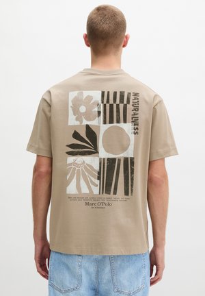 Mann trägt beige T-Shirt mit abstrakten, von der Natur inspirierten Motiven und schwarzem Text, kombiniert mit hellblauen Jeans, steht mit dem Rücken zur Kamera vor einfarbigem Hintergrund.